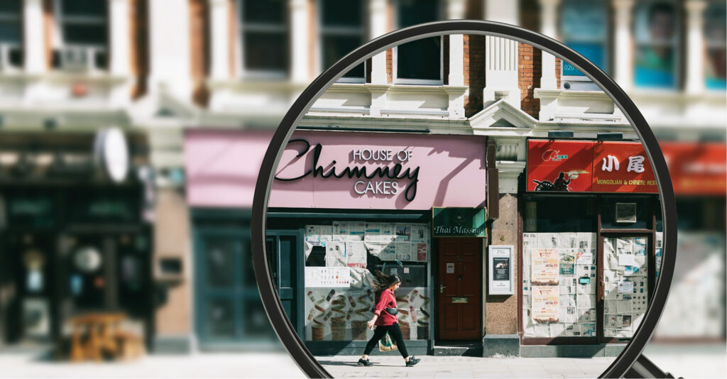 Magnifying glass highlighting a store on a street with multiple businesses, while a person walks by.